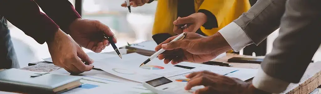 A group of people holding pens collaborates around a table with documents and charts spread out, discussing the costs involved in the website redesign project planned for 2024.
