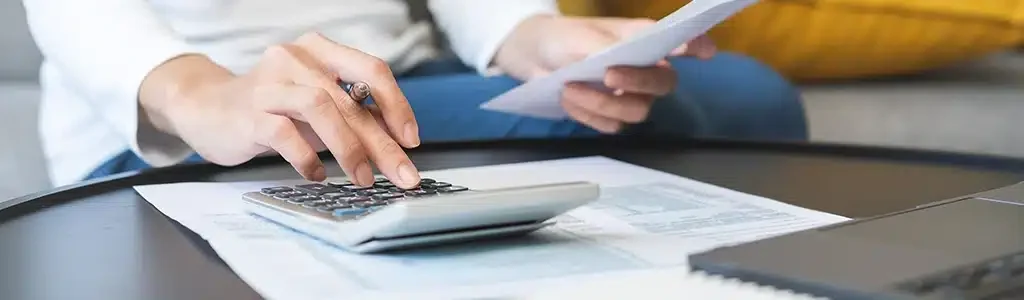 A person using a calculator and holding a piece of paper at a desk, with documents and a laptop nearby, is calculating the 2024 website redesign costs.